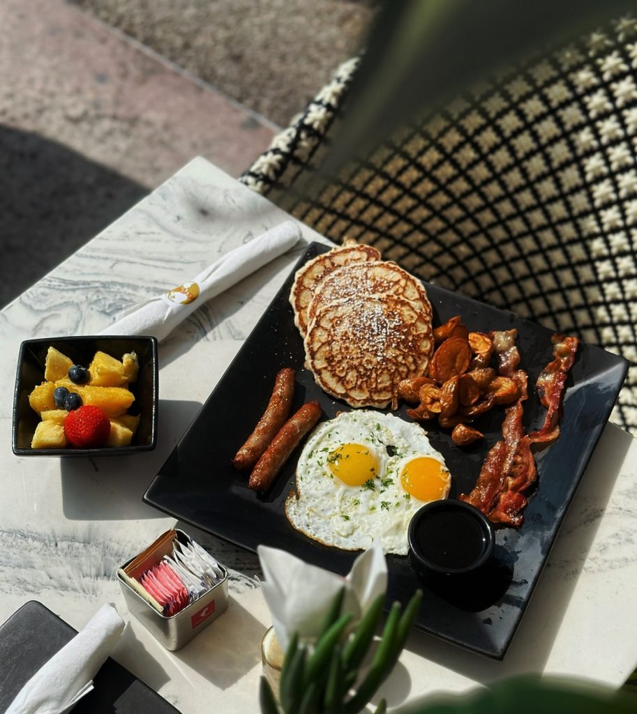 Breakfast at Montana’s Restaurant in Miami Beach featuring pancakes, sunny-side-up eggs, bacon, sausages, roasted potatoes, fresh fruit, and coffee served on a stylish outdoor patio table.
