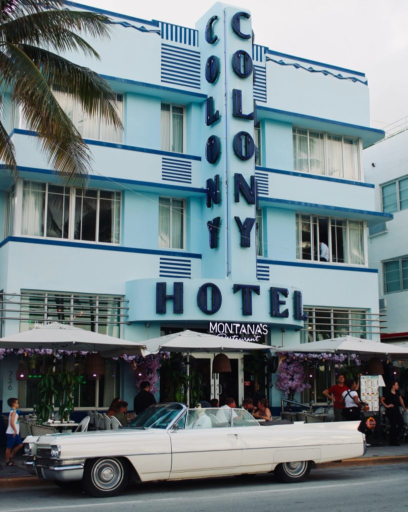 Exterior view of the iconic Colony Hotel on Collins Avenue in Miami Beach, home to Montana’s Restaurant, with its famous Art Deco blue facade and a classic vintage white car parked in front.
